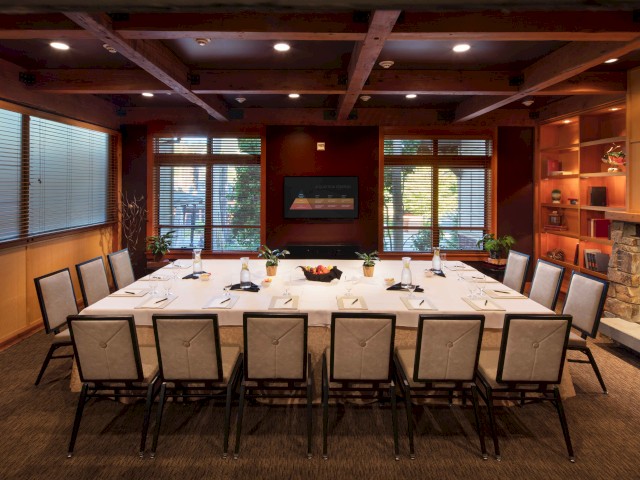 A conference room with a large U-shaped table, chairs, a fireplace, wooden beams, windows, and a TV on the wall.