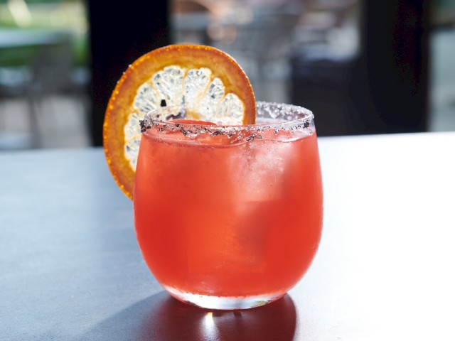 A vibrant red cocktail garnished with a lemon slice, served in a rounded glass with a salted rim, on a two-tone surface.