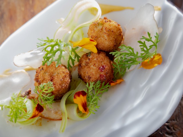 A plated gourmet dish features crispy fried ball appetizer garnished with fresh herbs, edible flowers, and creamy white sauce.