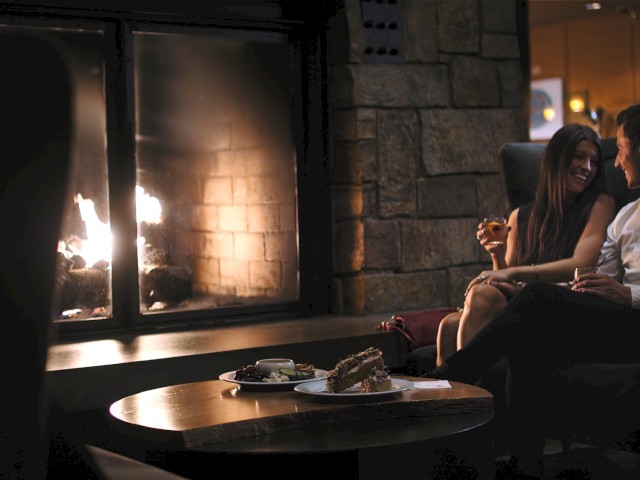 A couple enjoys drinks and dessert by a cozy fireplace in a warmly lit lounge area.