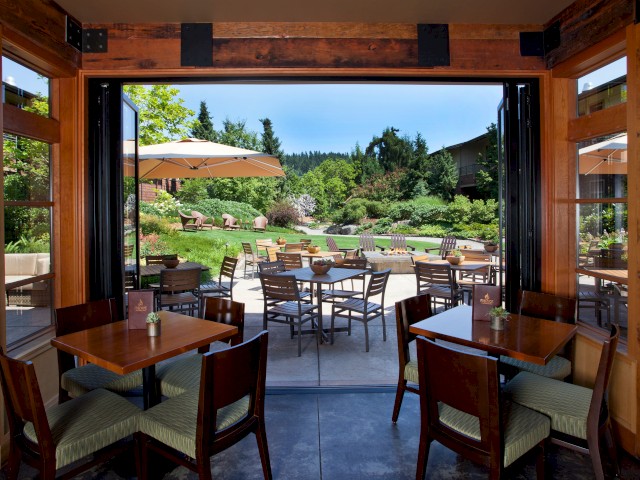 A cozy indoor dining area opens to a lush outdoor patio with tables, chairs, umbrellas, and vibrant greenery in perfect weather.