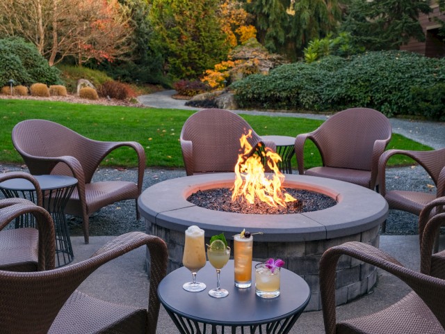An outdoor patio with a fire pit surrounded by chairs, and three tropical cocktails on a small table in front, in a lush garden setting.
