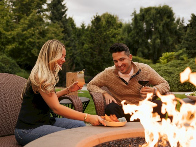 A woman and man enjoy drinks and food by a firepit in a lush, green outdoor setting, sharing a joyful moment together.