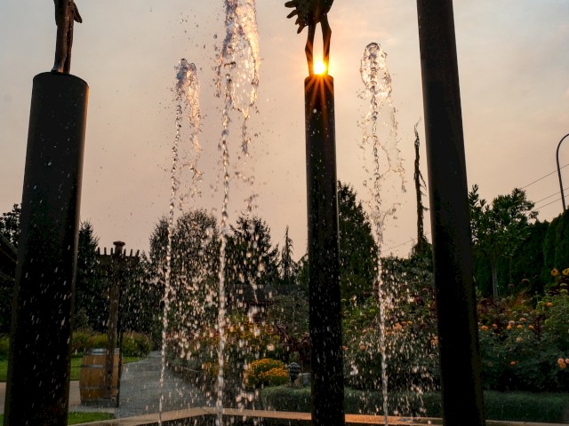 Fountain sculptures with water jets and metal figures in a garden at sunset.