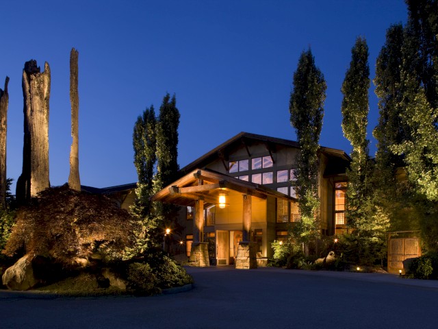 A beautiful modern house at dusk is illuminated by warm lights, with tall trees and a well-maintained landscape surrounding it.
