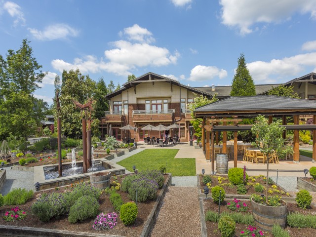 A beautifully landscaped garden with a fountain, patio seating, and a house in the background under a partly cloudy sky.