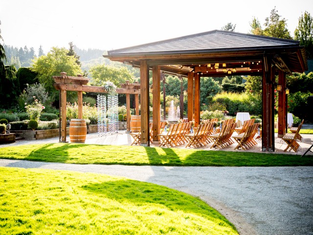 An outdoor event space with wooden chairs, tables, a pavilion, lush garden, and fountain, set in a scenic and sunny natural environment.