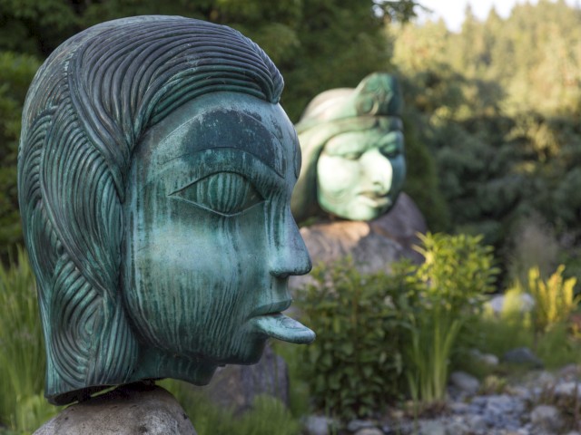 Two large, greenish-blue sculptures of human faces with closed eyes, surrounded by greenery and rocks.