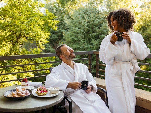 Two people in bathrobes enjoy morning tea and desserts on a balcony surrounded by greenery.