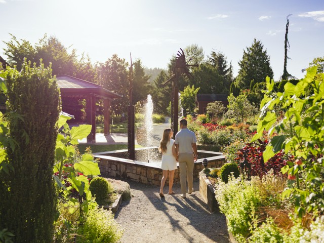 A couple walks hand-in-hand along a garden path near a fountain, surrounded by lush greenery and colorful flowers on a sunny day.