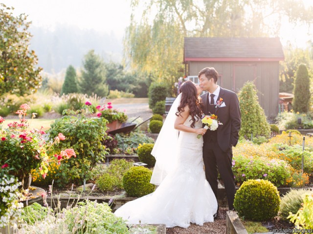 A bride and groom share a kiss in a sunny garden filled with colorful flowers and lush greenery, creating a romantic wedding scene.