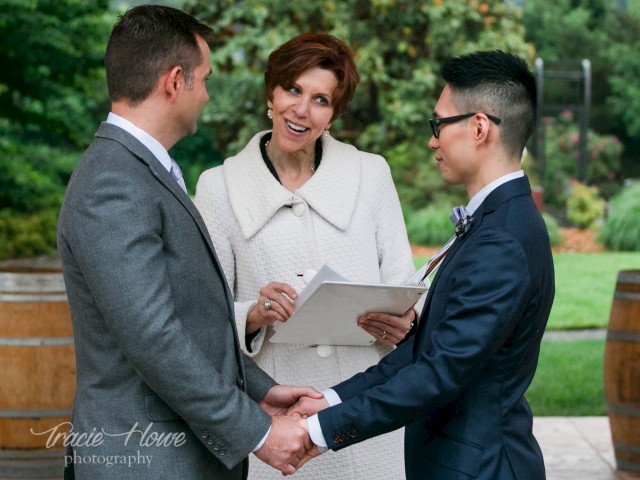 A woman officiates a same-sex wedding outdoors, during which a woman and a man exchange vows while holding hands.