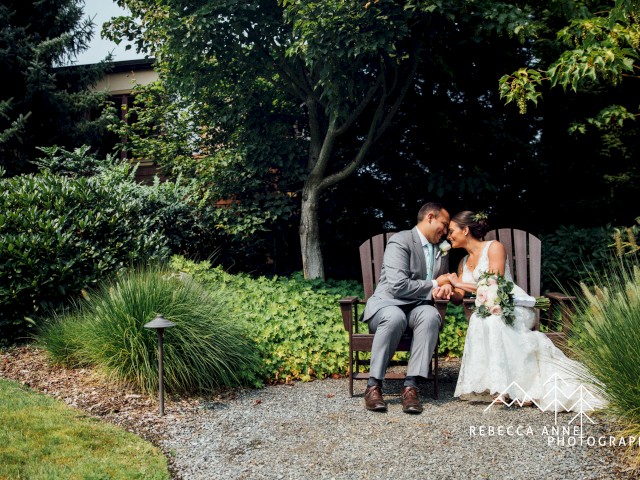 A bride and groom sit on a bench in a garden, sharing a happy, intimate moment surrounded by lush greenery during their wedding.
