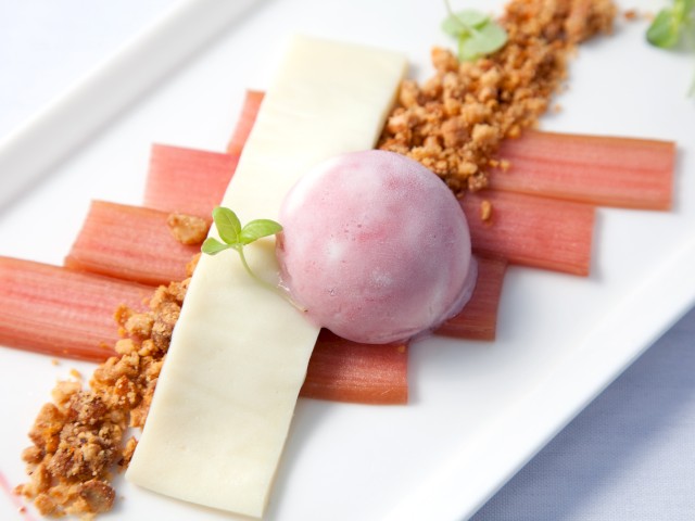 A plated dessert features pink rhubarb pieces, white chocolate, crumbly topping, a pink sorbet, and garnished with microgreens.