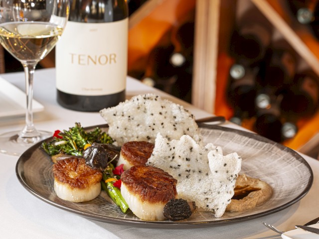 A gourmet dish with seared scallops, asparagus, black truffles, and crispy rice crackers, served with white wine and wine cellar in background.