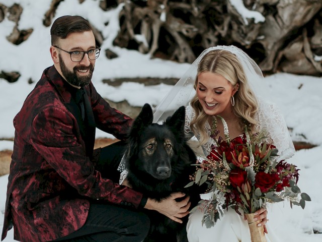 A bride and groom pose with their dog in a snowy outdoor setting during their wedding day.