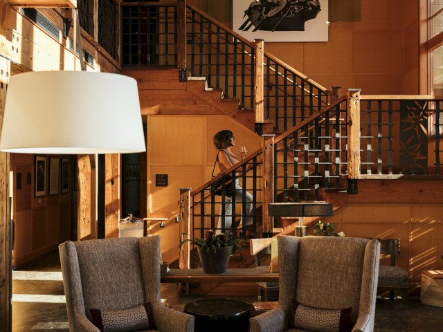A woman with curly hair is sitting on a staircase, holding a glass of wine, in a warmly lit, cozy wooden interior space.