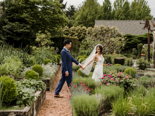 A bride and groom hold hands and walk through a lush garden with vibrant flowers, surrounded by greenery and whimsical sculptures.