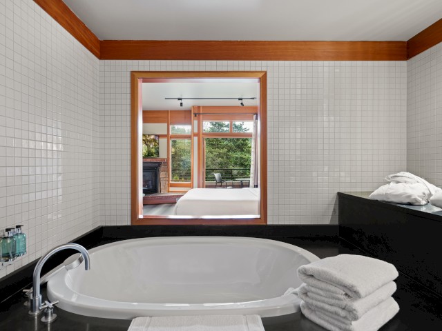A cozy bathroom with a view of a bedroom and balcony outside, featuring a large bathtub, neatly folded towels, and wooden accents.
