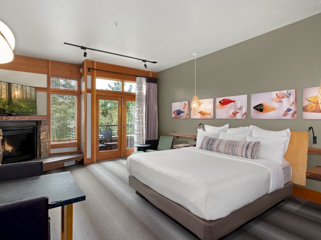 A cozy hotel room with a fireplace, large bed, artwork, and a balcony overlooking nature.