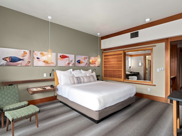 A hotel room featuring a large bed, wall art, seating, a bathroom visible through a connecting window, and modern decor elements.