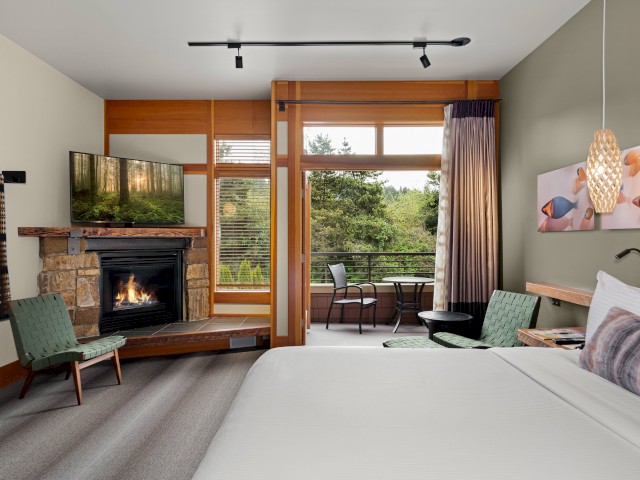 A cozy hotel room features a fireplace, a mounted TV, a bed, and a balcony with outdoor seating overlooking greenery.