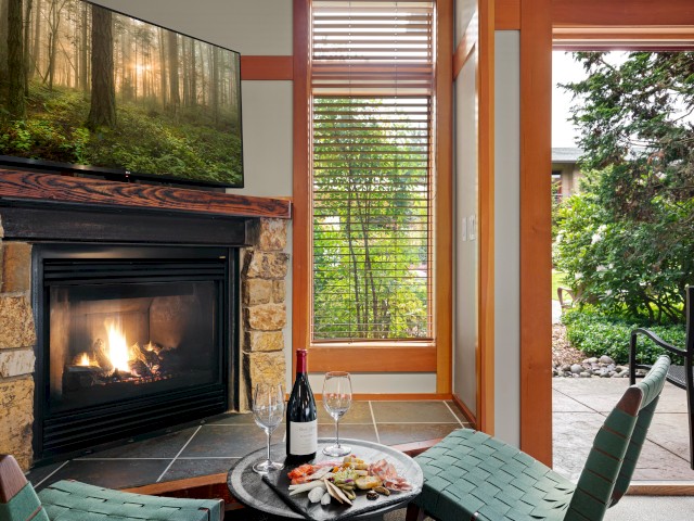 A cozy living room with a lit fireplace, a TV, a tray of snacks and wine, and a door opening to a lush green garden.