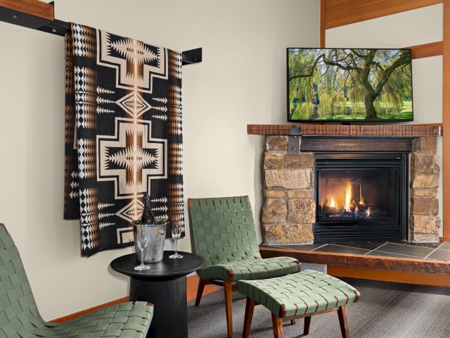 A cozy living room features a stone fireplace with a fire, a wall-mounted TV, green woven chairs, a small black table, and a colorful textile wall hanging.