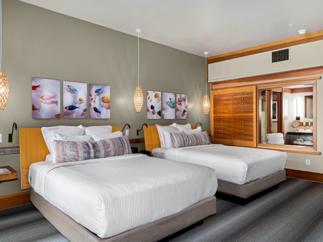 A hotel room with two beds, colorful artwork, hanging lights, and a large window looking into the bathroom area.