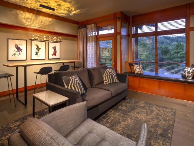 A cozy living room with a gray sofa, large windows, wooden accents, and modern artwork in a warm, inviting atmosphere.