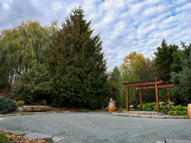 A peaceful garden with a gravel area, lush trees, a wooden bench, barrels, and a sculpture under a cloudy sky.
