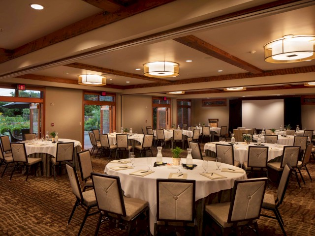 A spacious, well-lit banquet room set up with round tables, white tablecloths, chairs, and centerpieces for an event or gathering.
