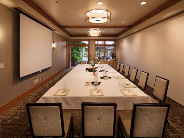 A well-set conference or meeting room with a long table, chairs, notepads, glasses, and a screen for presentations.