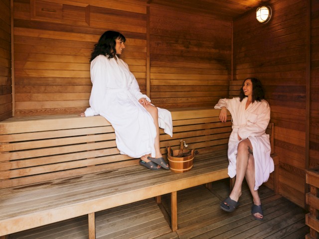Two women are relaxing in a wooden sauna, wearing robes and smiling at each other.