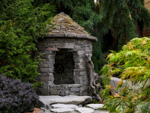A miniature stone shrine with a rounded roof, nestled among lush green plants and a stone pathway.
