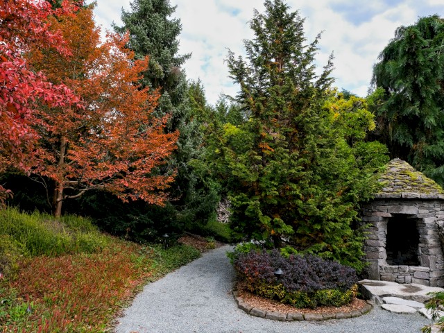 A peaceful garden scene features colorful autumn trees, a gravel path, and a small stone structure under partly cloudy skies.