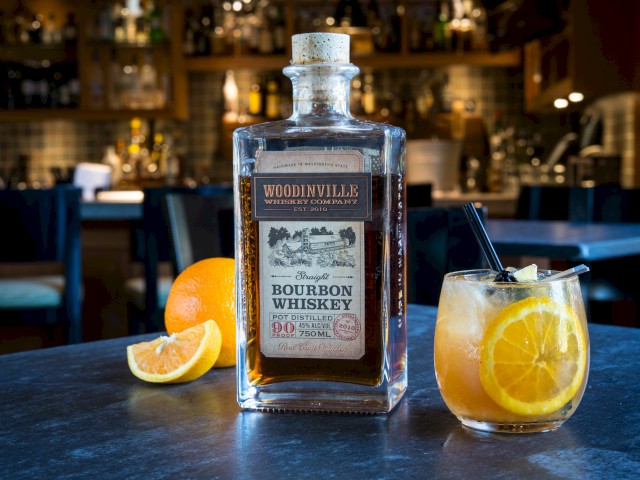 A bottle of Woodinville bourbon whiskey, an orange, and a citrus cocktail on a dark bar counter in a cozy bar setting.