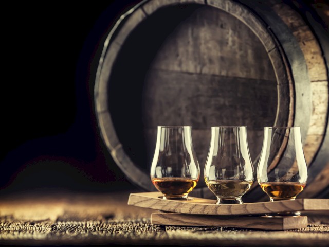 Three glasses of whiskey on a wooden tray in front of a large wooden barrel, with a dark background.