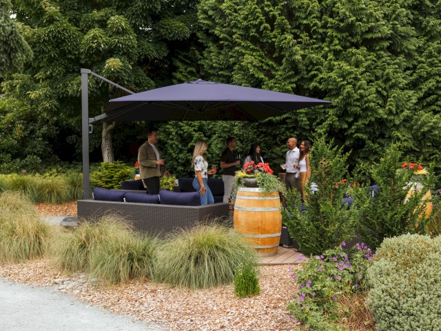 People are socializing outdoors on a patio with plants, a large umbrella, and a barrel used as a flower container.