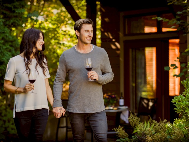 A couple holds hands and glasses of red wine in a cozy outdoor garden setting during the daytime.
