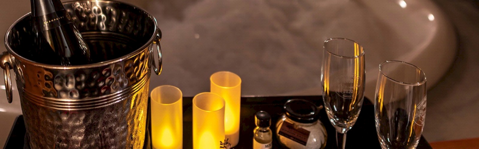 The image shows a romantic setup with a bubble bath, champagne in an ice bucket, candles, champagne glasses, and various spa items on a tray.