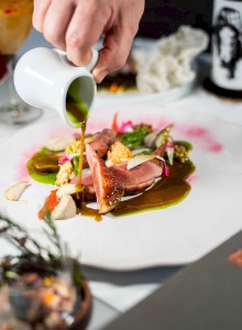 A hand pours sauce over an artfully arranged dish with meat and vegetables on a white plate.