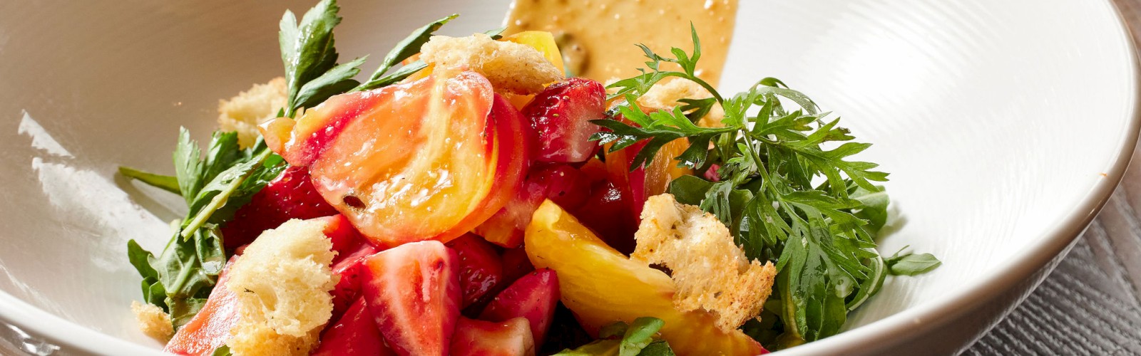 A bowl of fresh salad with tomatoes, greens, croutons, and a creamy dressing on the side, garnished with herbs, placed on a wooden surface.