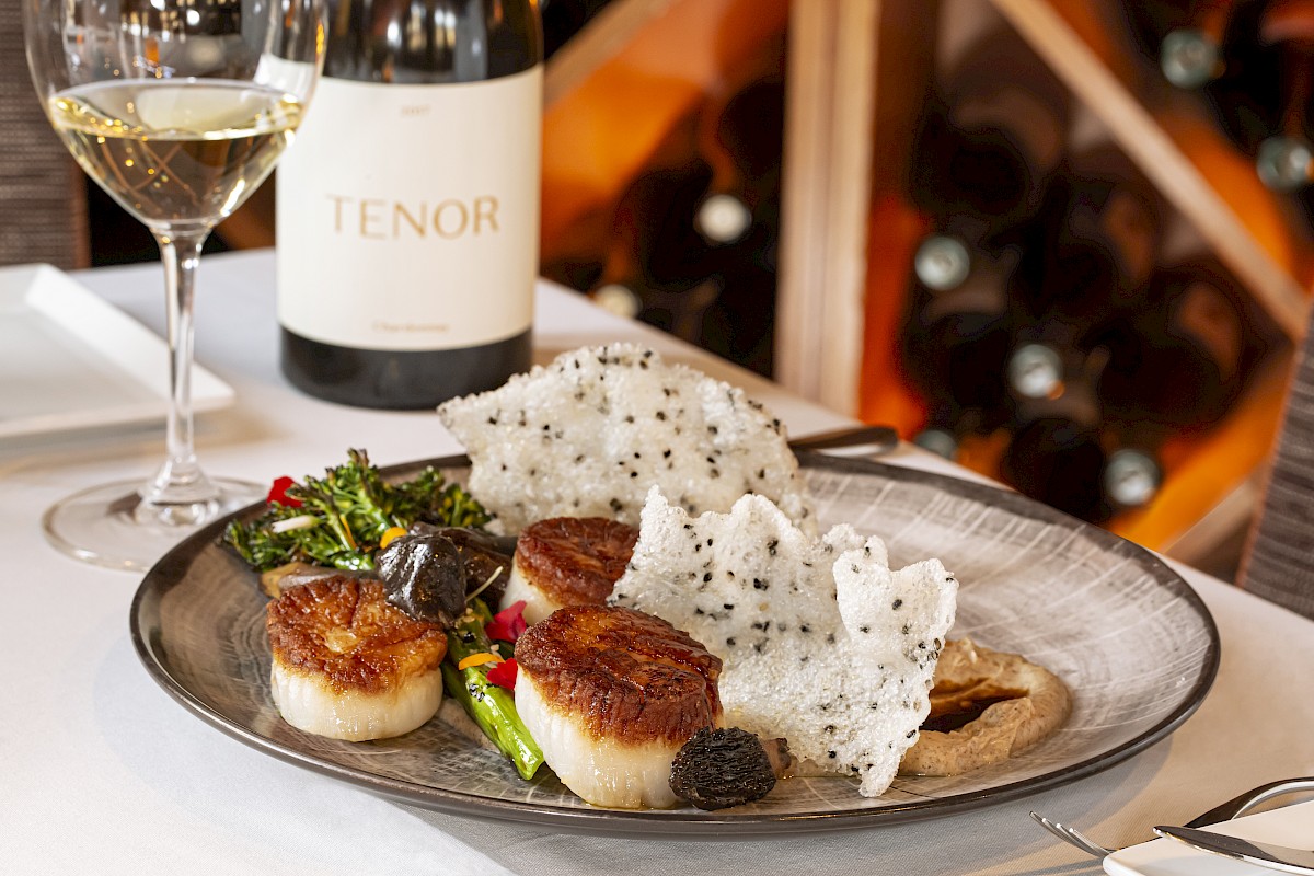 A gourmet meal with scallops, greens, and garnishes on a plate, accompanied by a glass of white wine and a bottle labeled