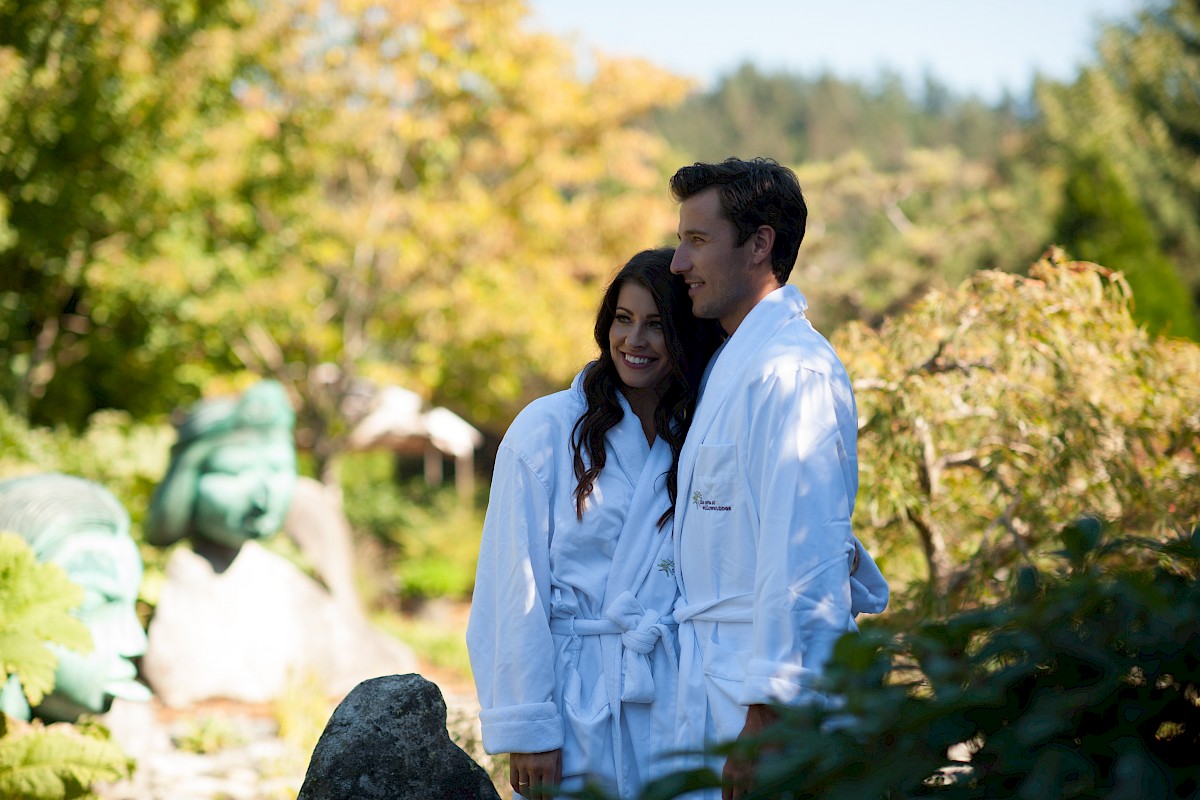A couple in white robes smiles while standing outdoors in a lush garden. They appear relaxed, enjoying the beautiful scenery around them.
