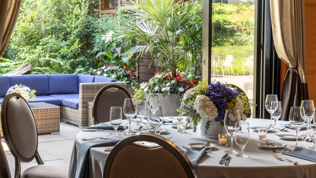 A beautifully set dining table indoors with elegant glassware and floral arrangements, overlooking a lush garden patio.