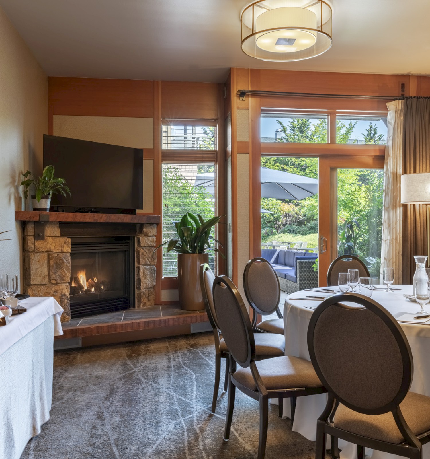 A cozy dining room with a round table set for a meal, a fire in the fireplace, a TV, plants, and a view of a garden outside.