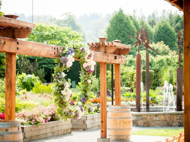 A garden wedding setup with wooden arches, floral wreaths, barrels, and a fountain, surrounded by lush greenery and sunny ambiance.