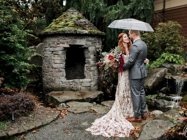 A woman in white dress and veil holds an umbrella, standing next to a stone house with lush greenery in the background.