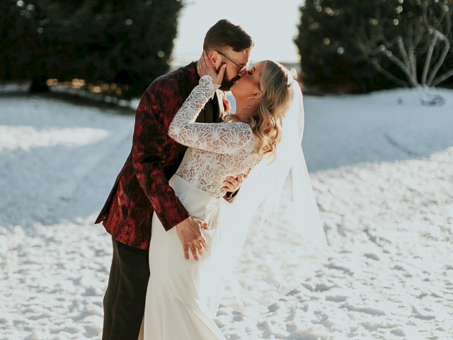A man and a woman in wedding attire kiss, surrounded by snowy landscape and trees